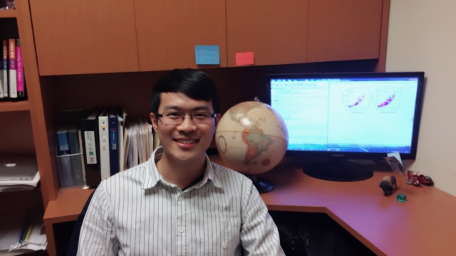 Dr. Youtong Zheng poses for the camera in front of his desk which has some books, a globe, and his computer