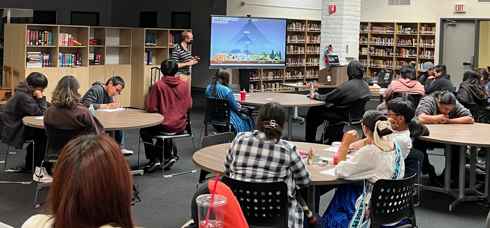 Feldman is at the front of a wide classroom, speaking in front of a television screen depicting a scientific graphic. The students are seated at circular tables distributed around the room and watching Feldman's presentation. 