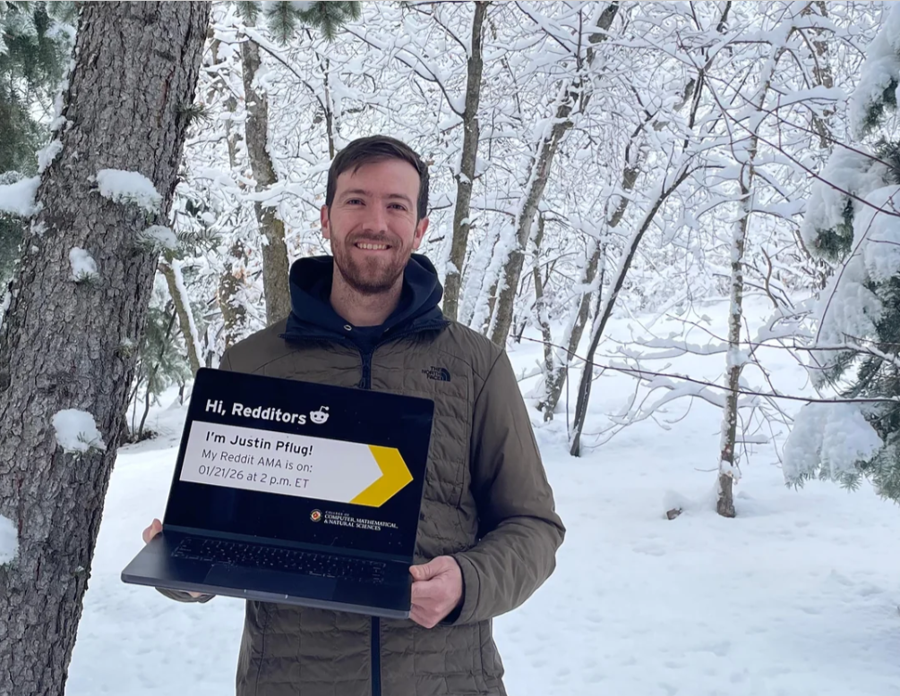 Justin stands in front of a wintery scene holding a laptop
