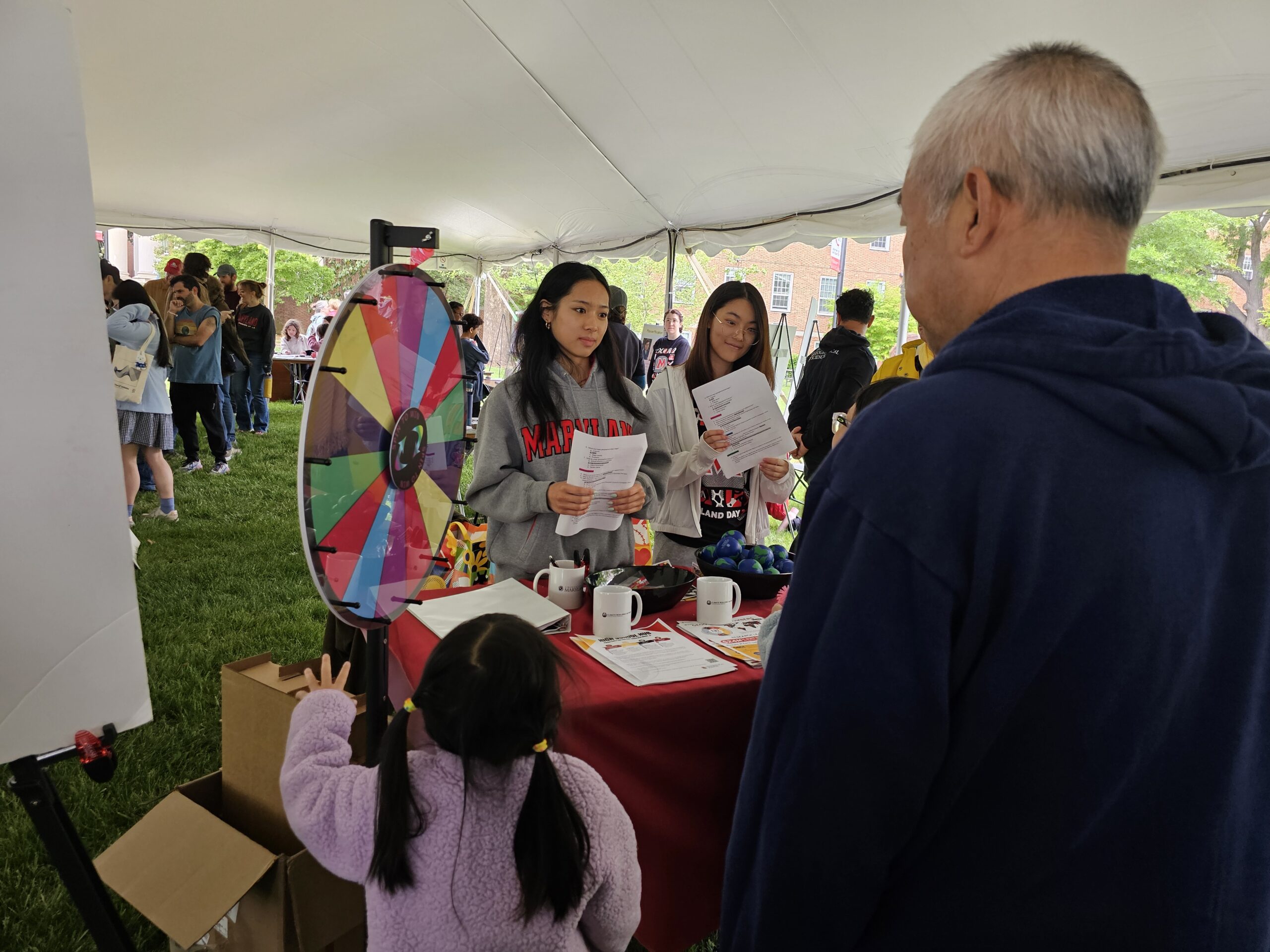 Geographical Sciences students standing besides a colorful spinning wheel and behind a red table laden with swag.