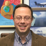 A man in businesswear and glasses smiles in front of a colorful background of posters