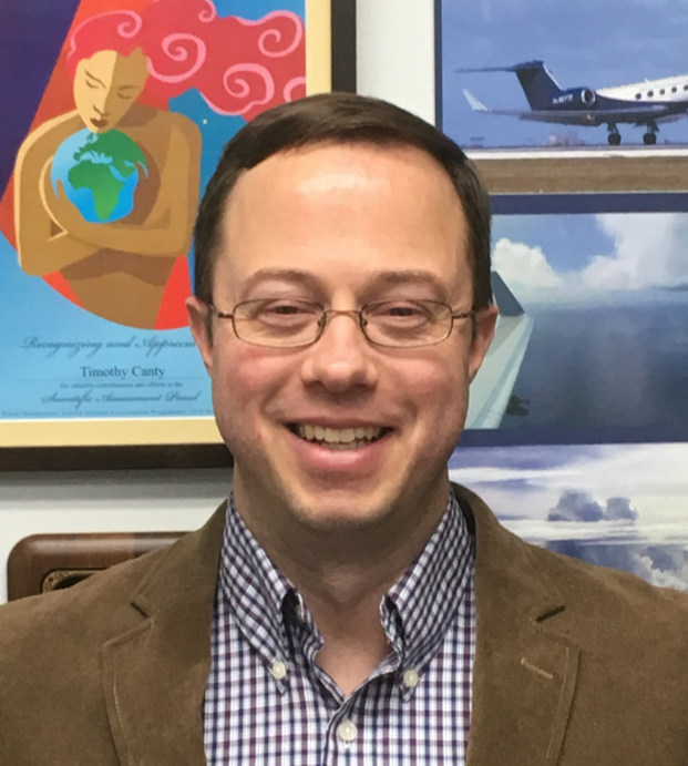 A man in businesswear and glasses smiles in front of a colorful background of posters