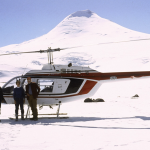 An aged photo of a helicopter in front of a snowy mountain. Three people pose in front of it.