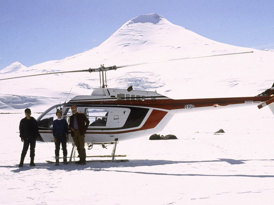 An aged photo of a helicopter in front of a snowy mountain. Three people pose in front of it.