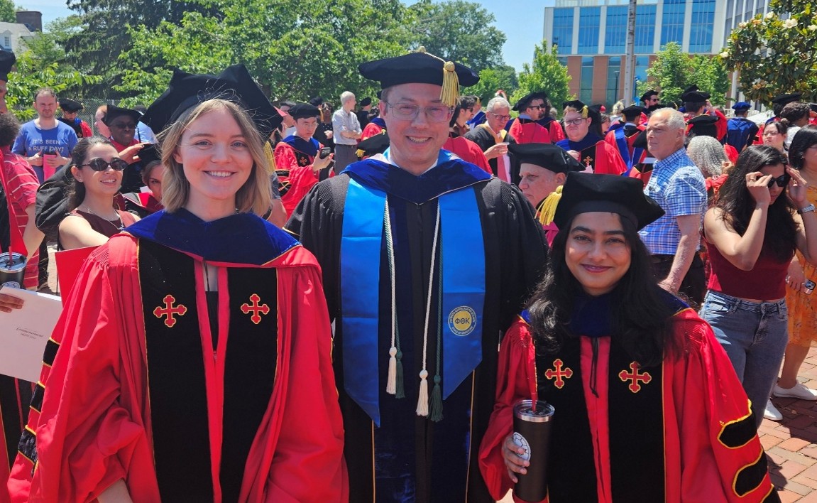 A professor at Commencement wearing blue regalia, alongside two new doctors wearing red regalia.